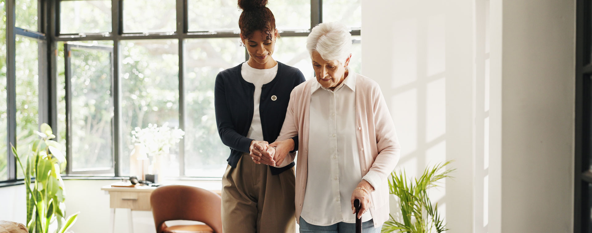 Seniorin mit Gehstock und Parkinson wird von einer unterstützenden Person im Alltag zuhause begleitet